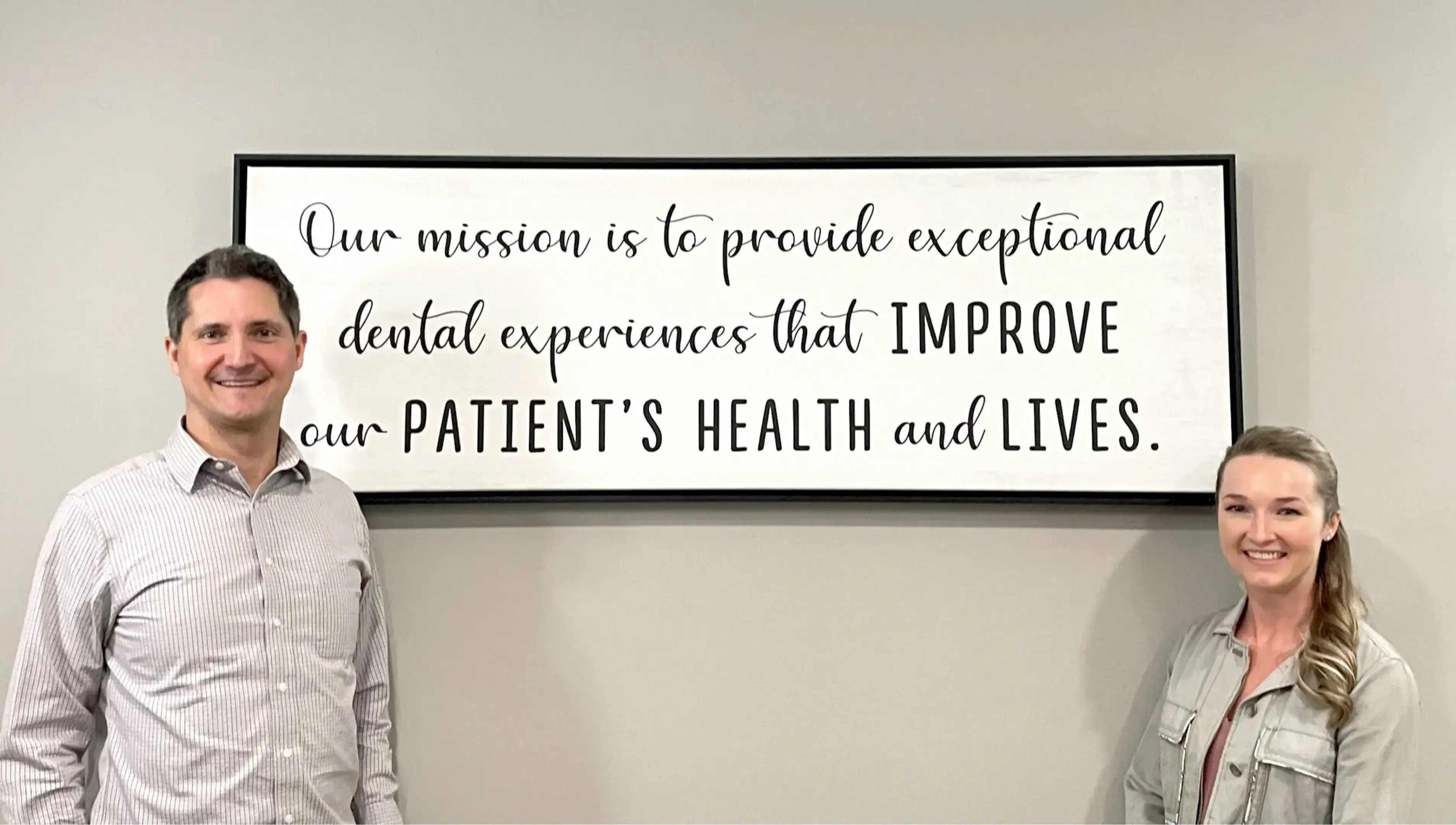 A man and woman standing and smiling in front of a wall sign that reads: 'Our mission is to provide exceptional dental experiences that improve our patient's health and lives.'